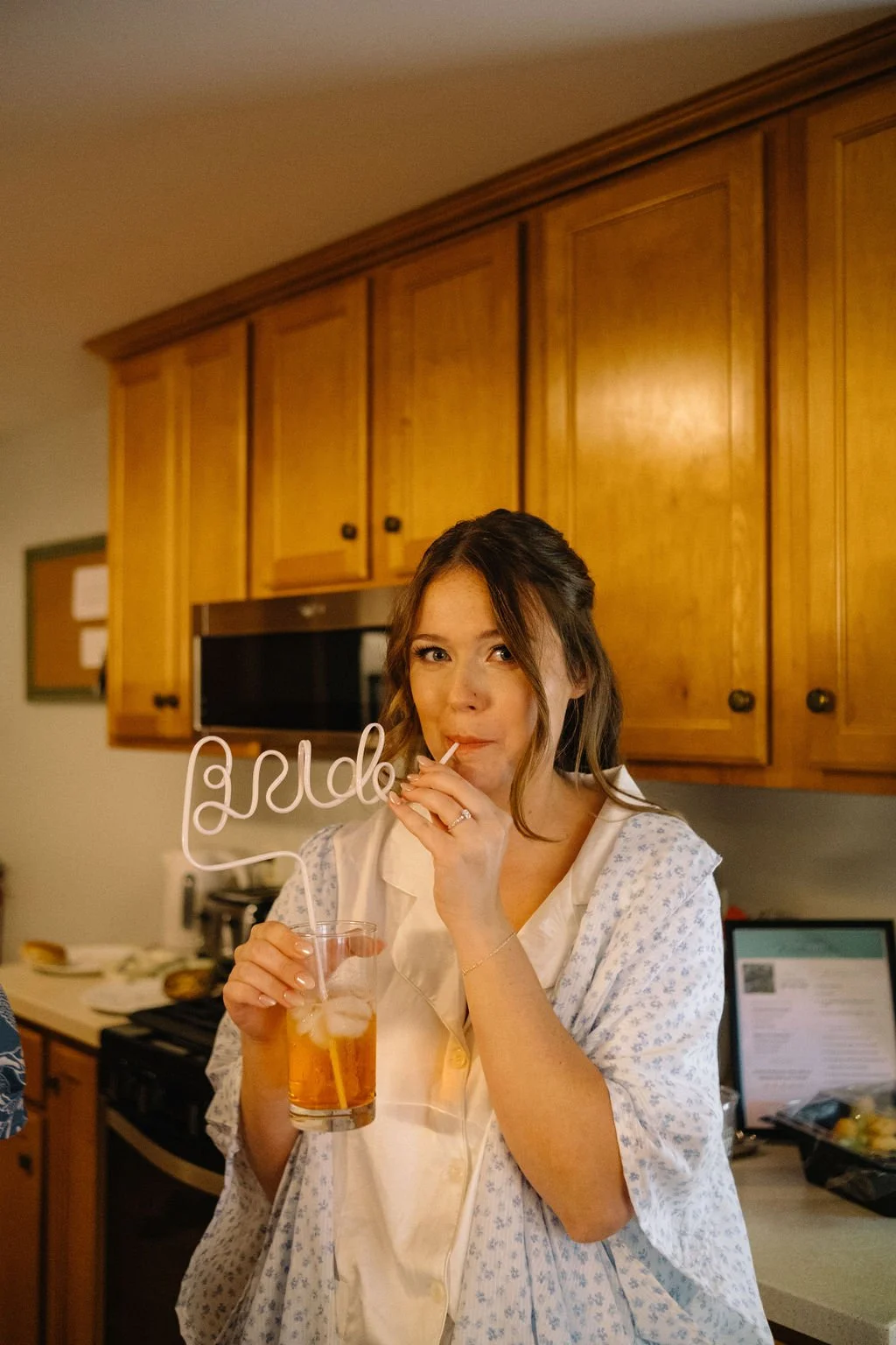 A woman in a hospital gown holding a drink with ice and a straw, drinking from a stirrer that spells 'bride', standing in a kitchen.