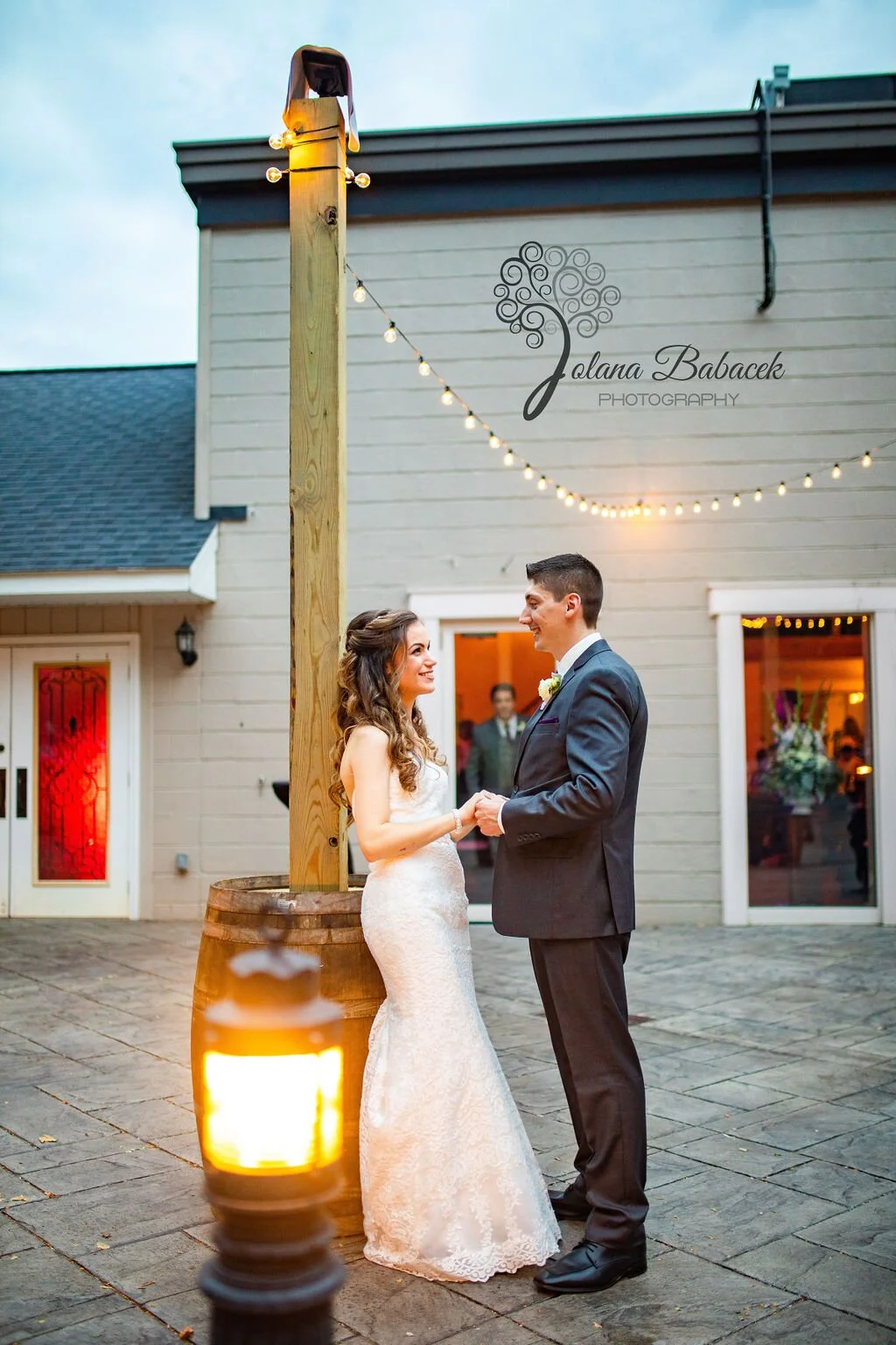 A bride and groom holding hands and smiling at each other during their wedding ceremony outdoors, with string lights overhead and a house in the background.