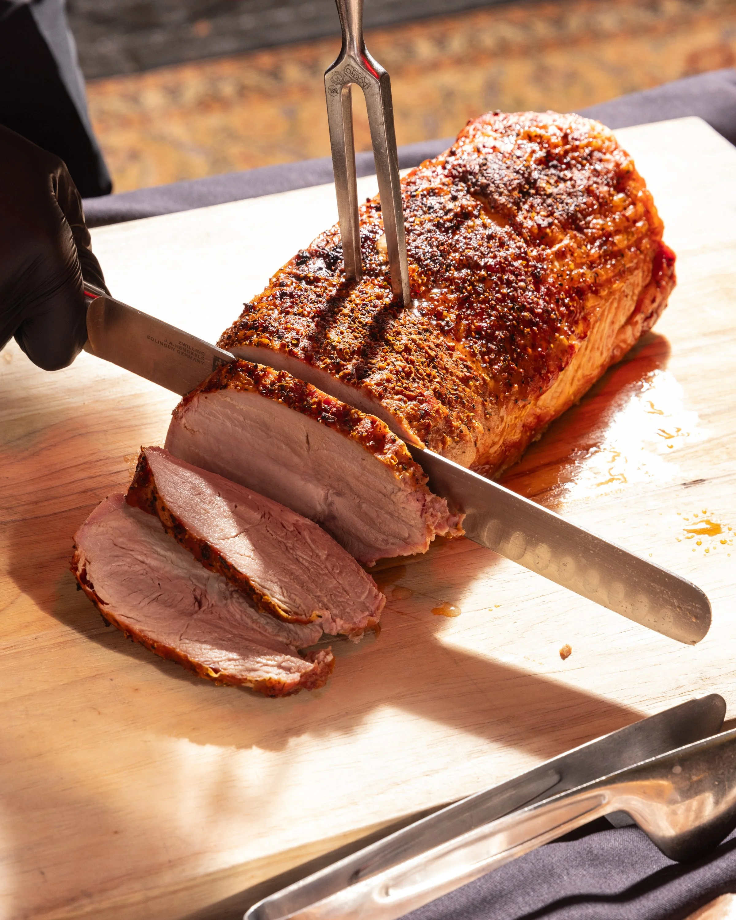 A chef in black gloves slices a roasted meat, likely pork, on a wooden cutting board using a large chef's knife. The meat has a seasoned crust and is being cut into slices, with a portion already sliced and laid out. The scene is set outdoors on a ta
