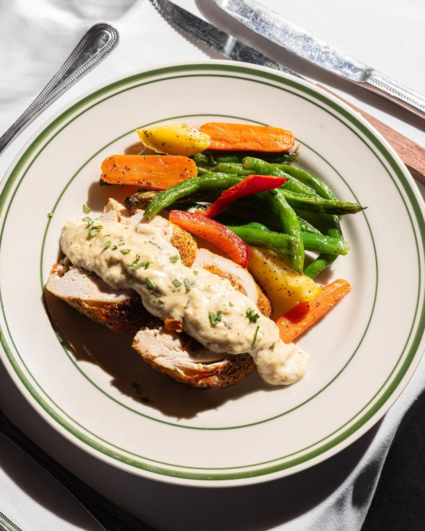 A plate with a serving of sliced turkey topped with a creamy white sauce, alongside roasted carrots, green beans, and potatoes, on a white plate with green stripes.