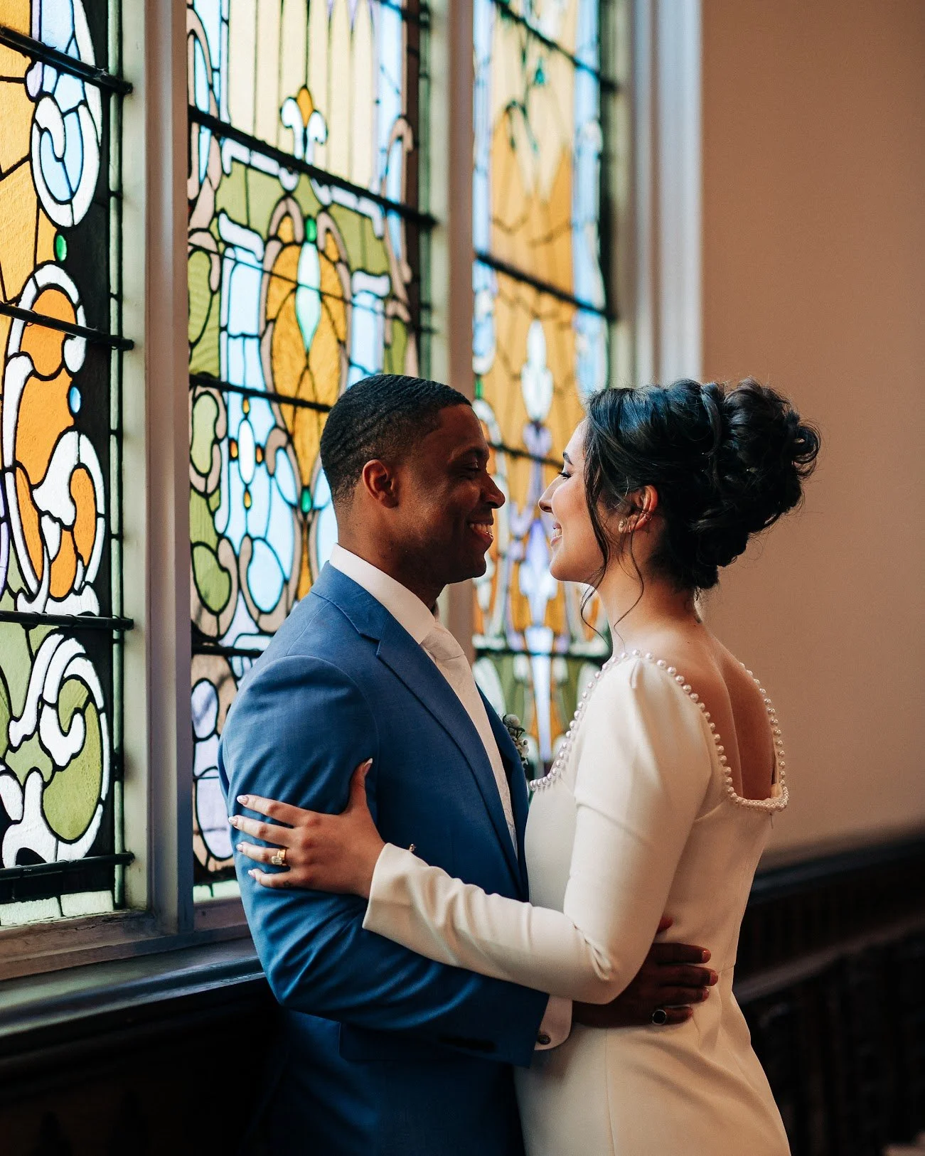 A newlywed couple with dark skin and curly hair in a church, standing close and smiling at each other, with a stained glass window in the background.