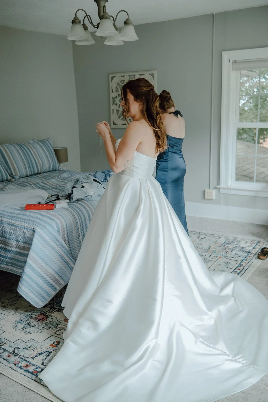 A bride in a white wedding dress holding a necklace, with another woman in a blue dress standing by a window in a bedroom.