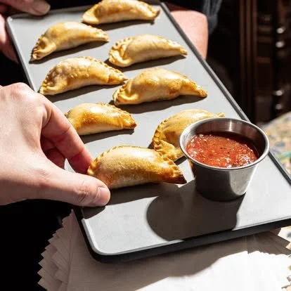 Hand holding a tray with ten fried empanadas and a small bowl of red dipping sauce.