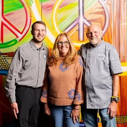 Three people standing in front of a colorful mural with peace symbol designs, smiling at the camera.