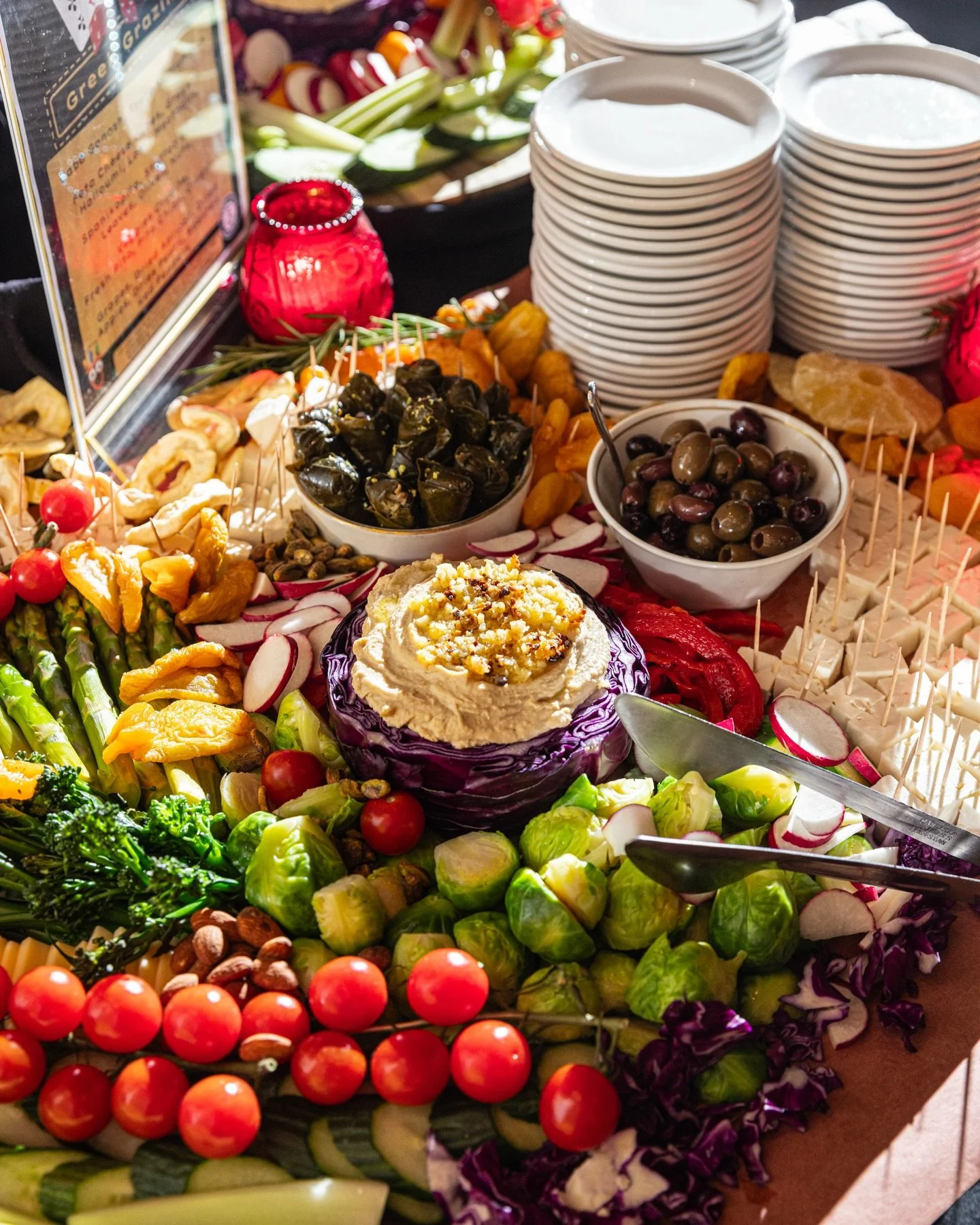 A colorful vegetable and snack platter featuring cherry tomatoes, Brussels sprouts, sliced radishes, grapes, olives, spinach, and hummus with sliced vegetables. There are stacks of white bowls, a serving spoon, and decorative elements in the backgrou