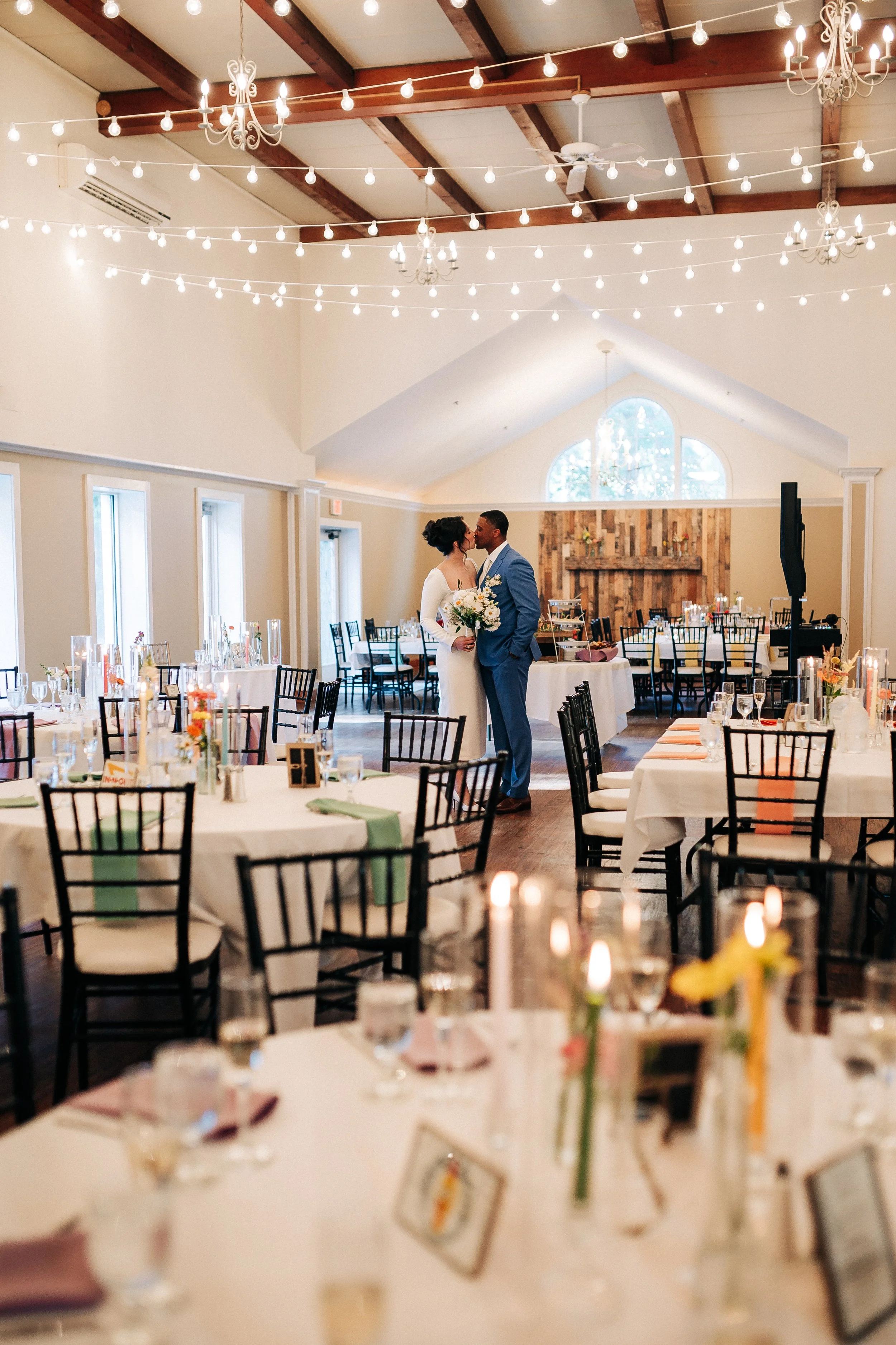 A wedding reception with a bride and groom kissing in the center of a decorated banquet hall, surrounded by round tables with floral centerpieces and candles, string lights hanging from the ceiling, and a rustic wooden wall in the background.