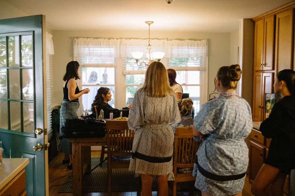 A group of women gathered in a kitchen with a wooden dining table, some standing and others sitting, in a well-lit room with large windows and white curtains.