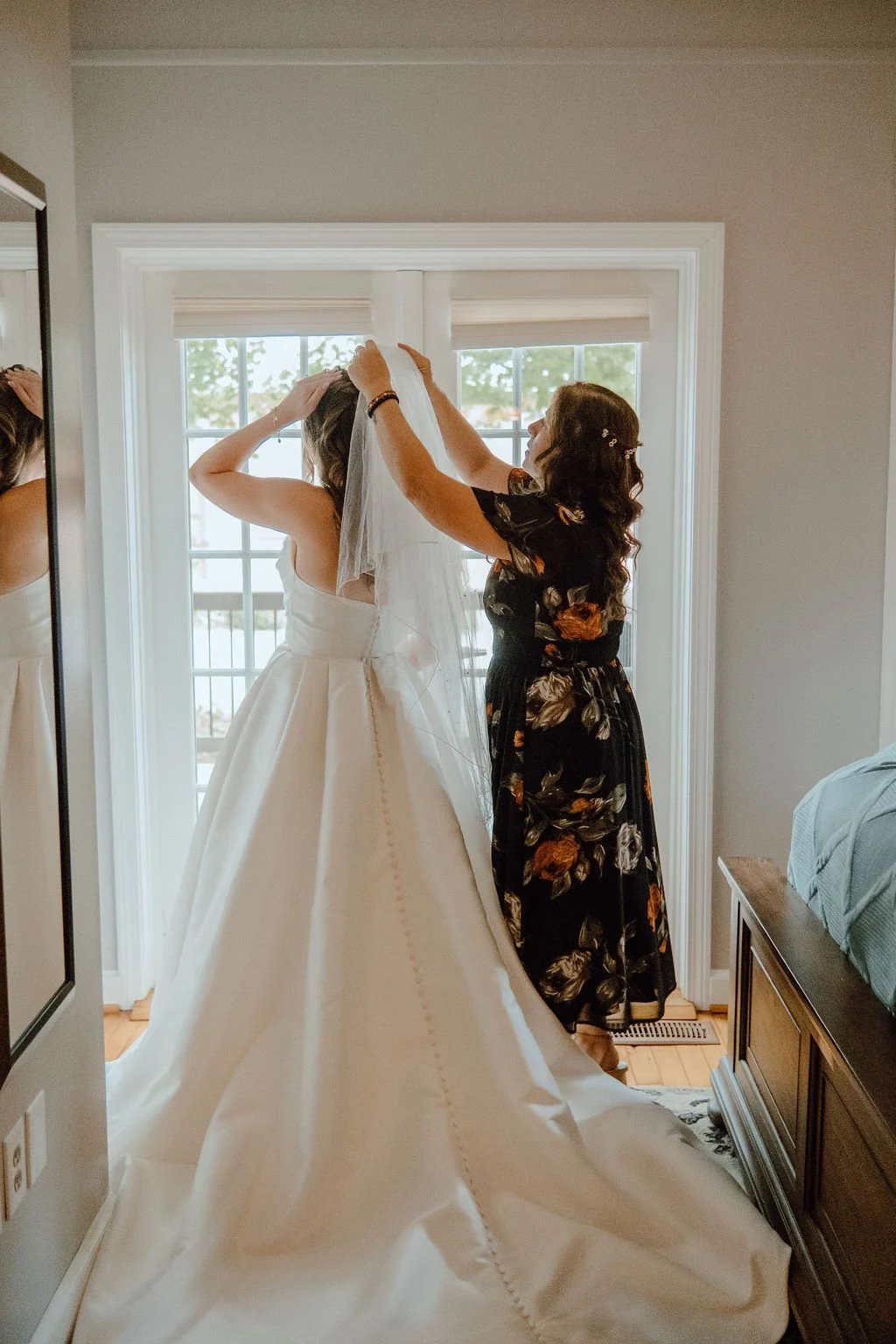 A woman helping a bride with her veil in a room with a large window.