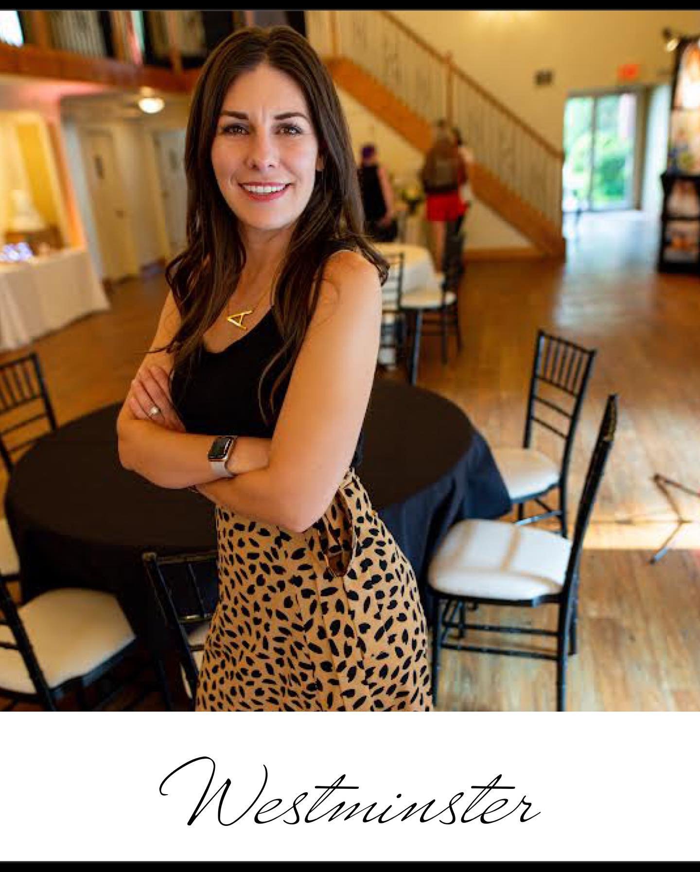 A smiling woman with dark brown hair wearing a black sleeveless top and a gold necklace, standing with crossed arms in a restaurant or cafe with wooden floors, chairs, and a staircase in the background.