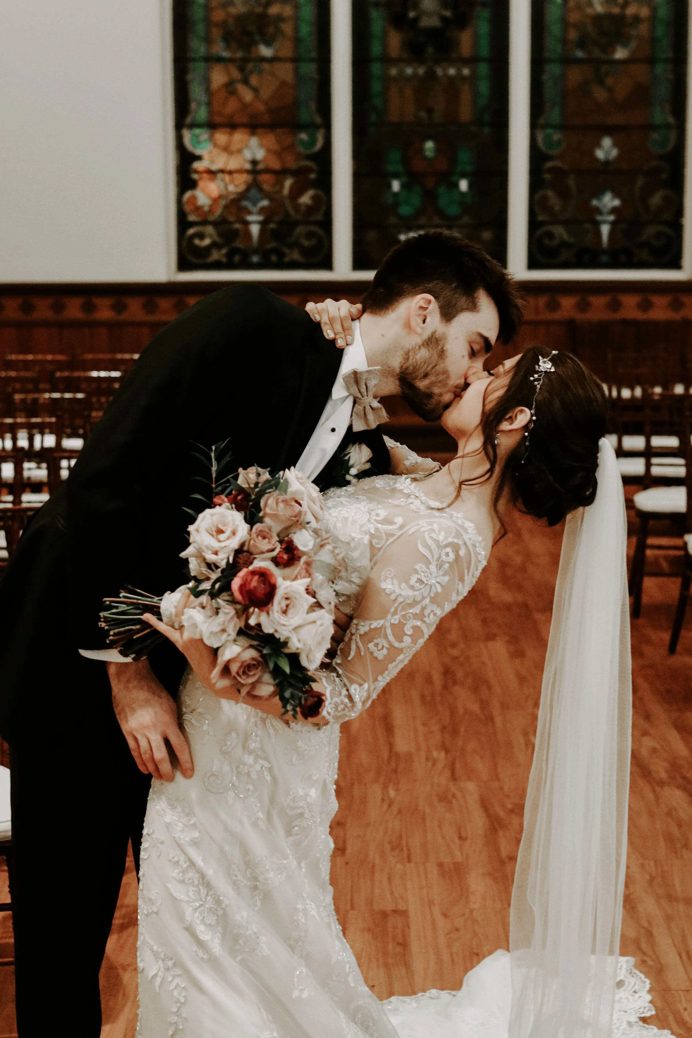 A bride and groom share a kiss during their wedding ceremony in a church, with wooden pews and stained glass windows in the background. The bride wears a lace wedding gown and a tiara, holding a bouquet of pink and red roses, while the groom wears a black tuxedo with a tan bow tie.