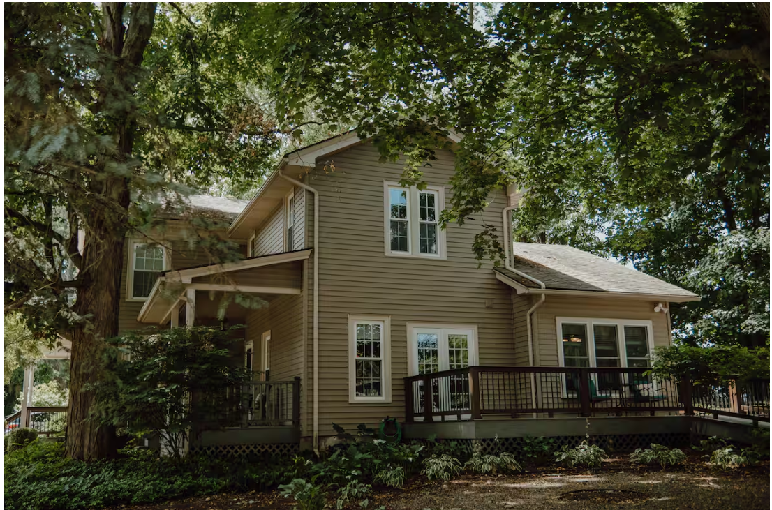 A two-story house with beige siding, multiple windows, and a wooden deck surrounded by green trees and landscaping.