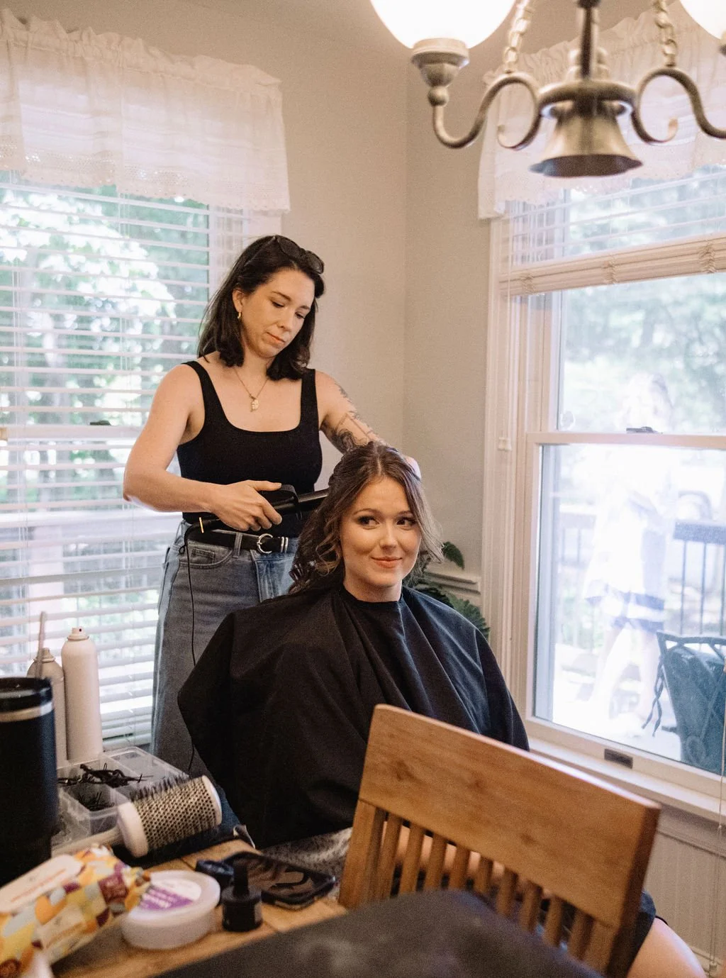 A woman with shoulder-length brown hair sitting in a chair and smiling while getting her hair styled by a hairstylist with dark hair in a sleeveless black top.