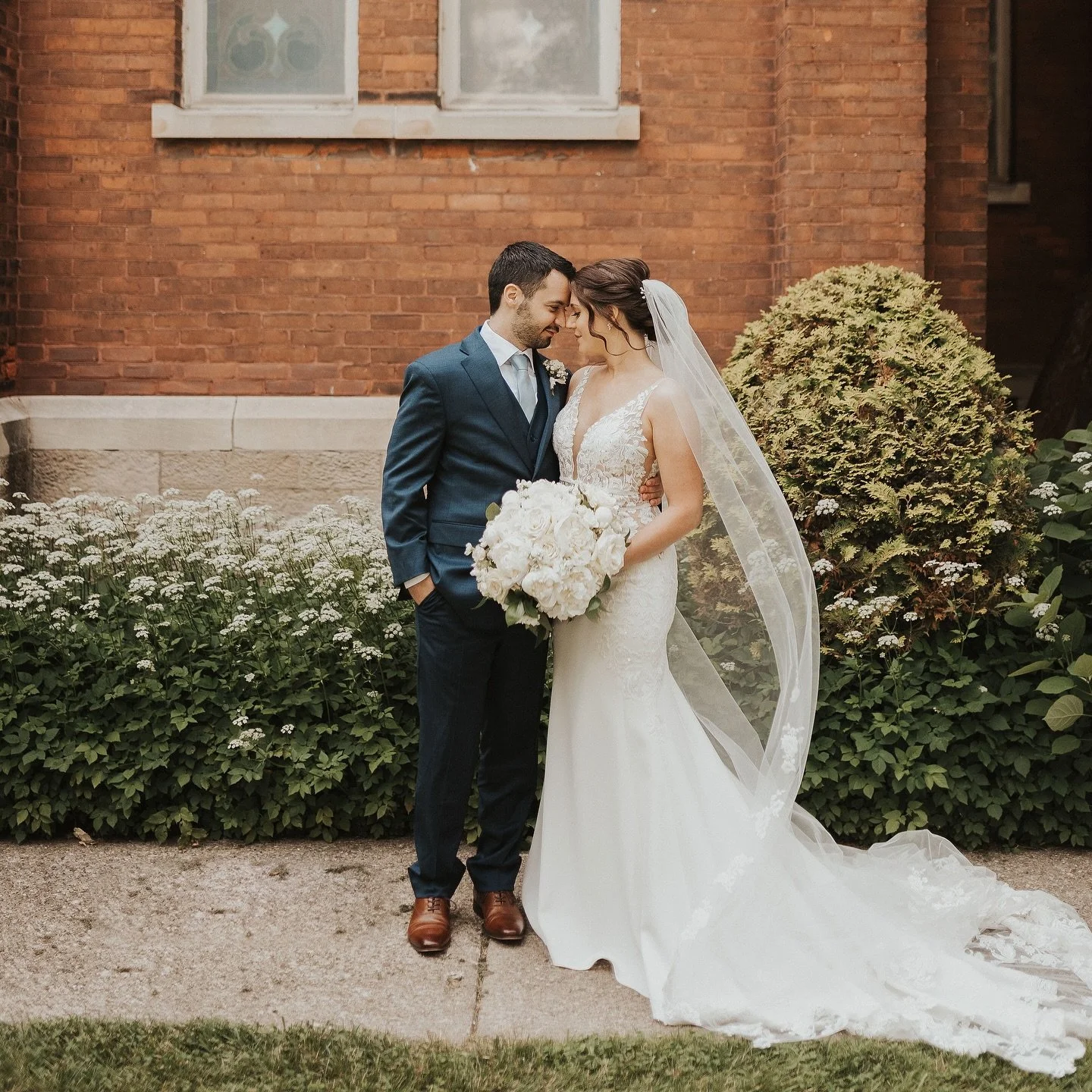 A bride and groom standing close together outdoors, touching foreheads, with the bride holding a large bouquet of white flowers. The bride wears a white lace wedding dress with a sheer veil, and the groom wears a dark blue suit, white shirt, and brown shoes. They stand in front of a brick building with greenery and white flowers.
