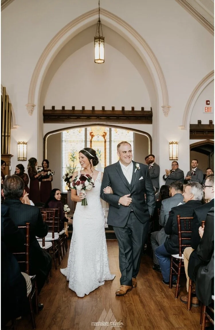 A bride and groom walking down the aisle at their wedding ceremony inside a church, surrounded by guests sitting in chairs and clapping.