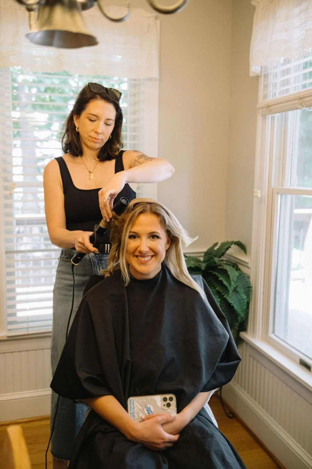 A woman with blonde hair receiving a haircut from a hairstylist in a room with large windows and a potted plant.