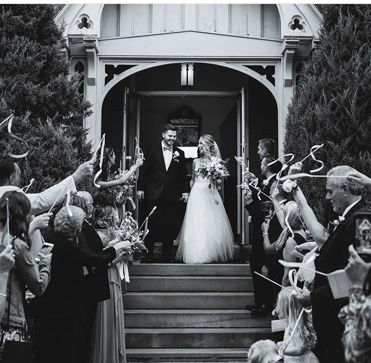 A wedding couple, a man in a tuxedo and a woman in a wedding dress, leaving a church while guests celebrate with waving ribbons and taking photos.