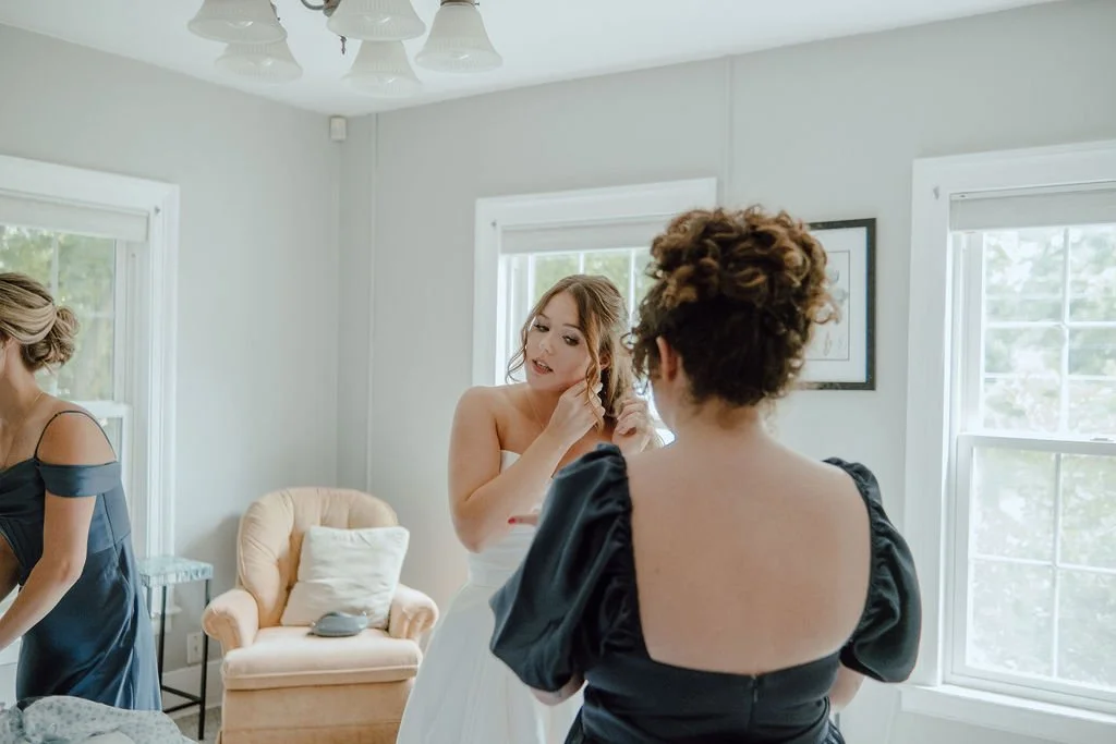 Two women in a bright, white room with large windows, one in a white dress and the other in a black dress, preparing for a wedding. An additional woman in a blue dress is partially visible, and a beige armchair with a pillow is in the background.