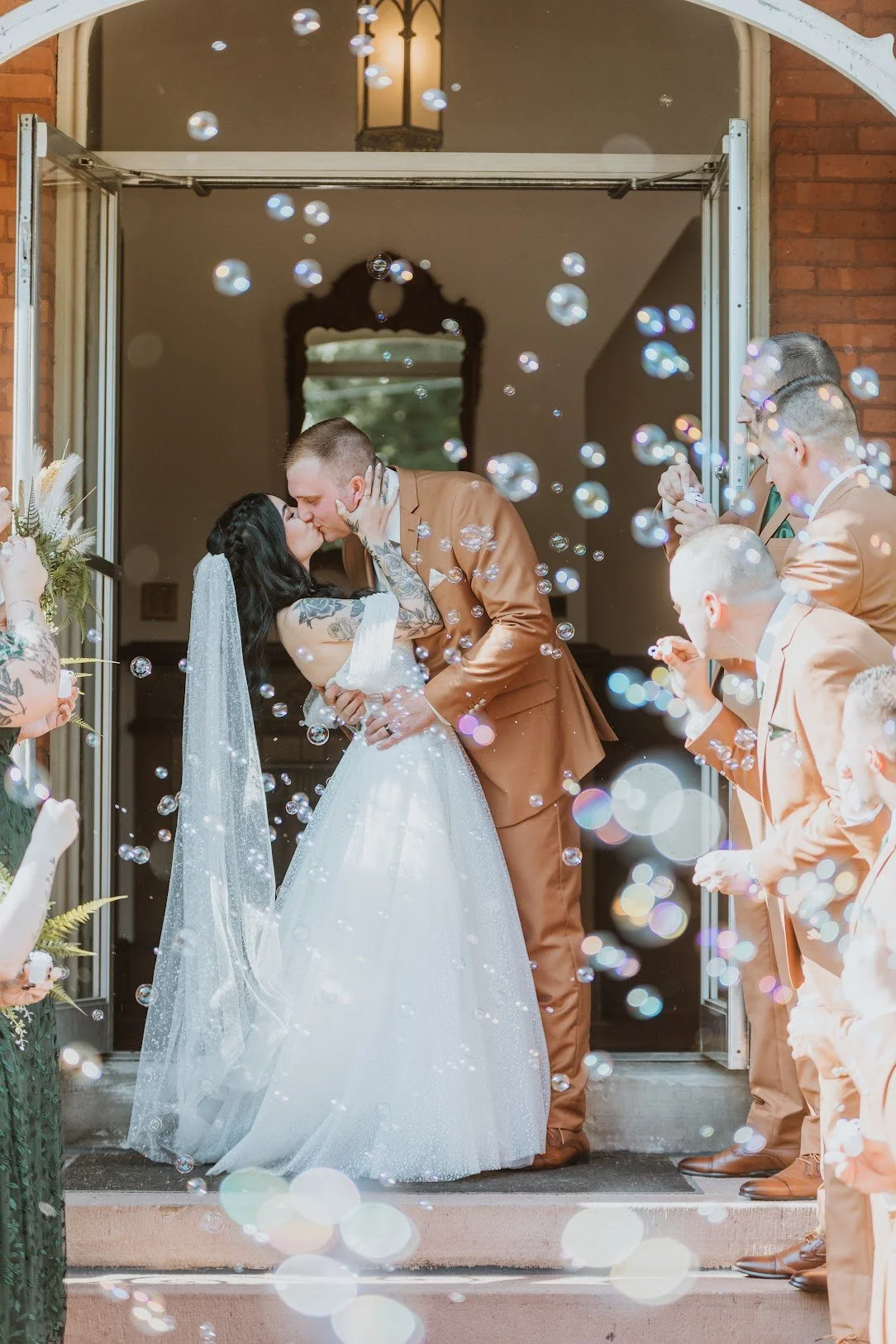 A newlywed couple sharing a kiss during their wedding ceremony as bubbles float around them, with friends and family on the porch celebrating.