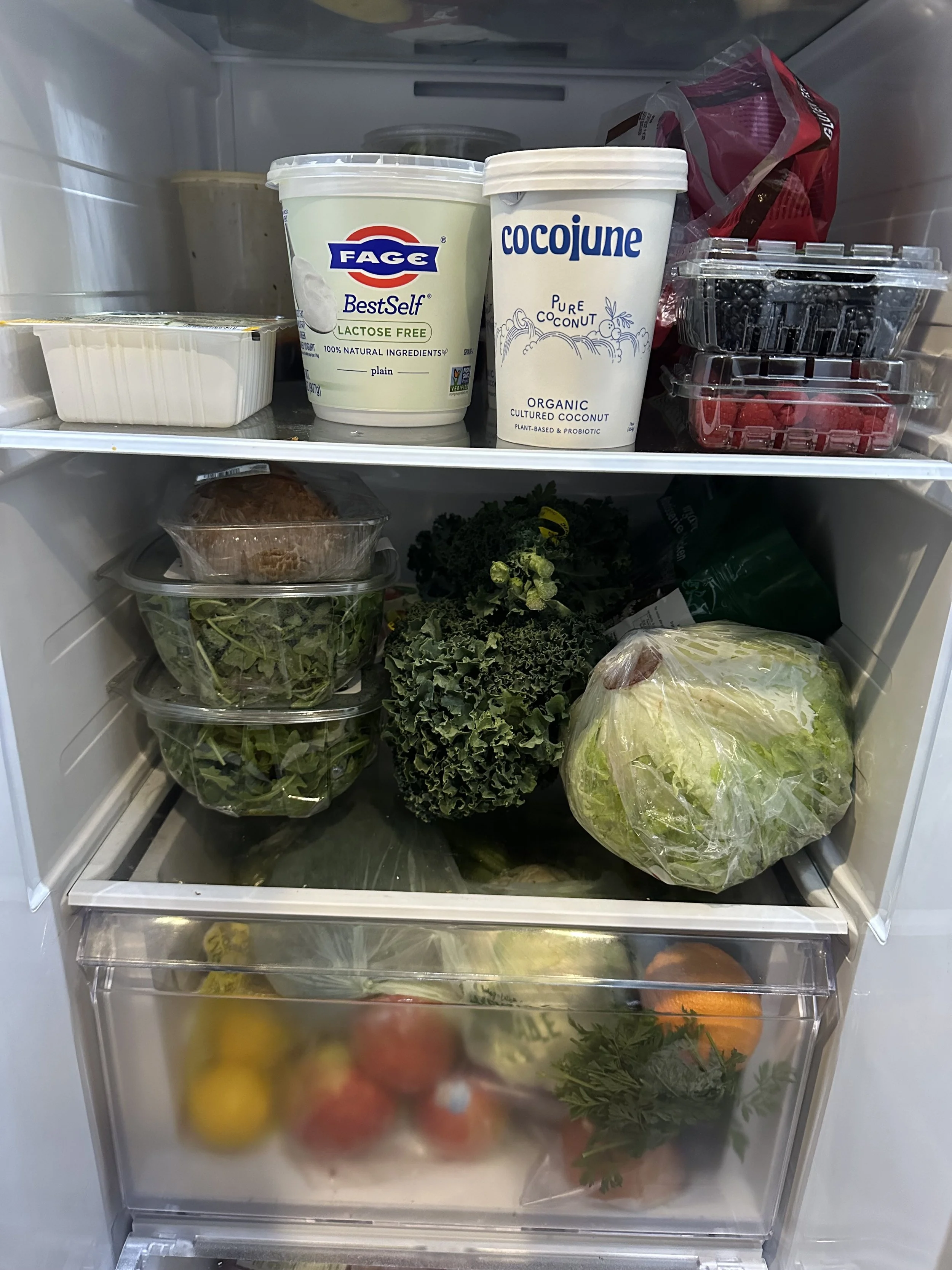 Inside a refrigerator with containers of yogurt, berries, greens, a head of lettuce, carrots, oranges, and various other fresh produce.