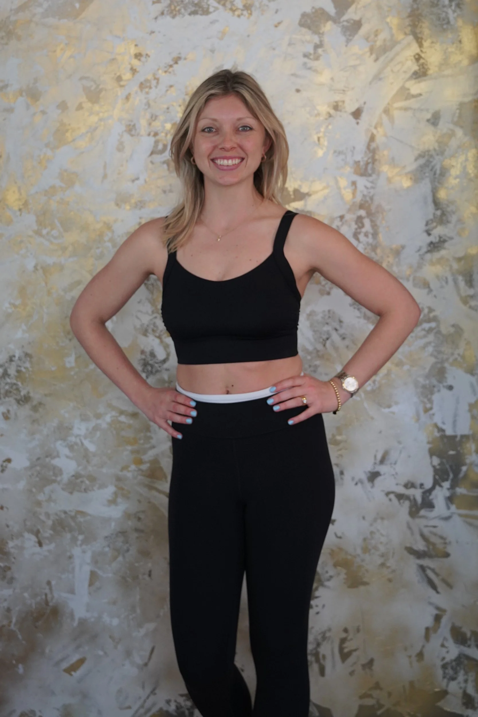 A woman with blonde hair smiling, wearing a black sports bra and black leggings, standing with her hands on her hips in front of a textured wall.