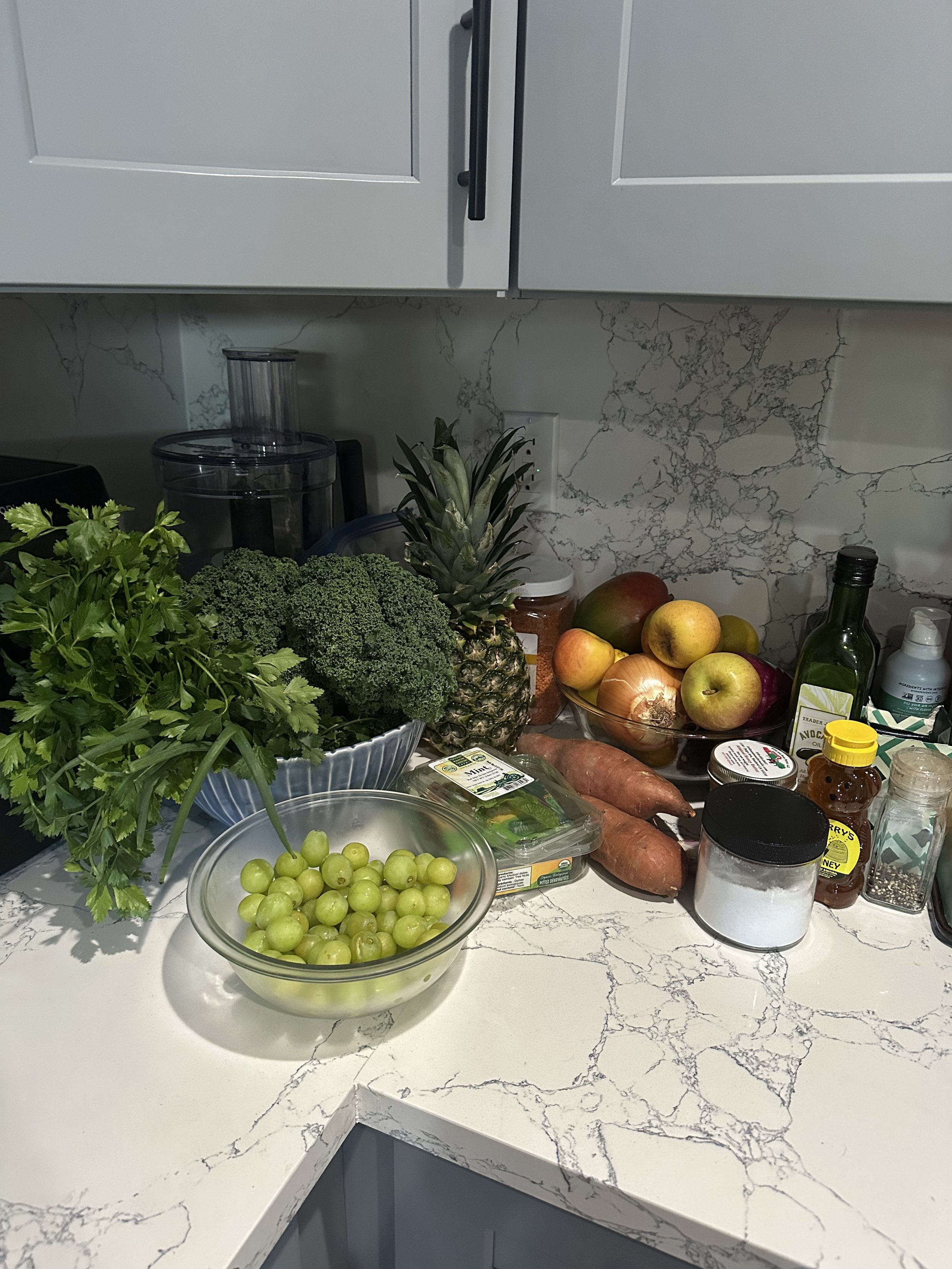 Kitchen counter with various fresh fruits and vegetables including green grapes, broccoli, pineapple, apples, sweet potatoes, and herbs, along with cooking ingredients like honey, salt, and oil.