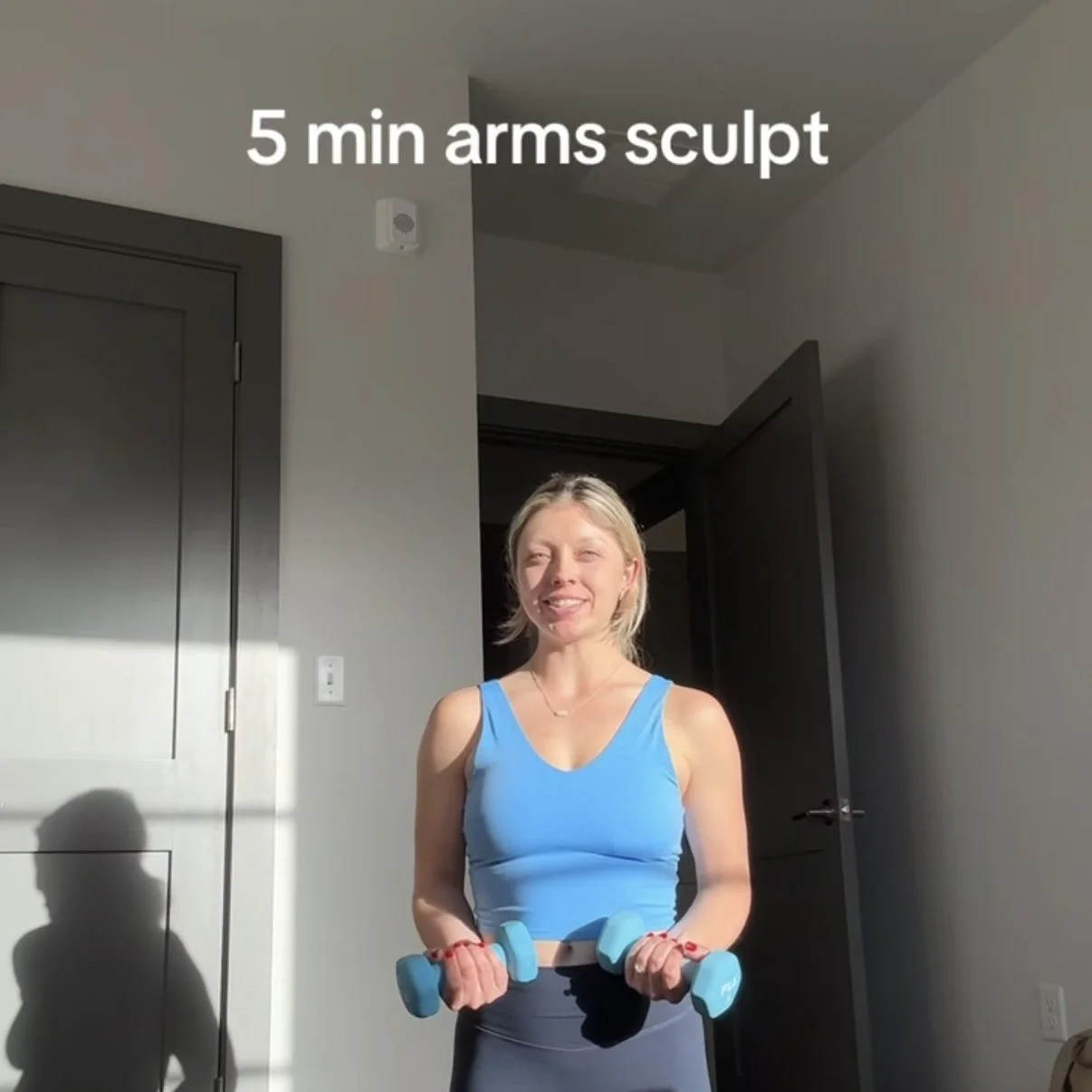 Woman in blue sports bra and black leggings performing a 5-minute arms sculpting exercise with light blue dumbbells in a room with gray walls and dark doors, sunlight casting shadows on the wall.