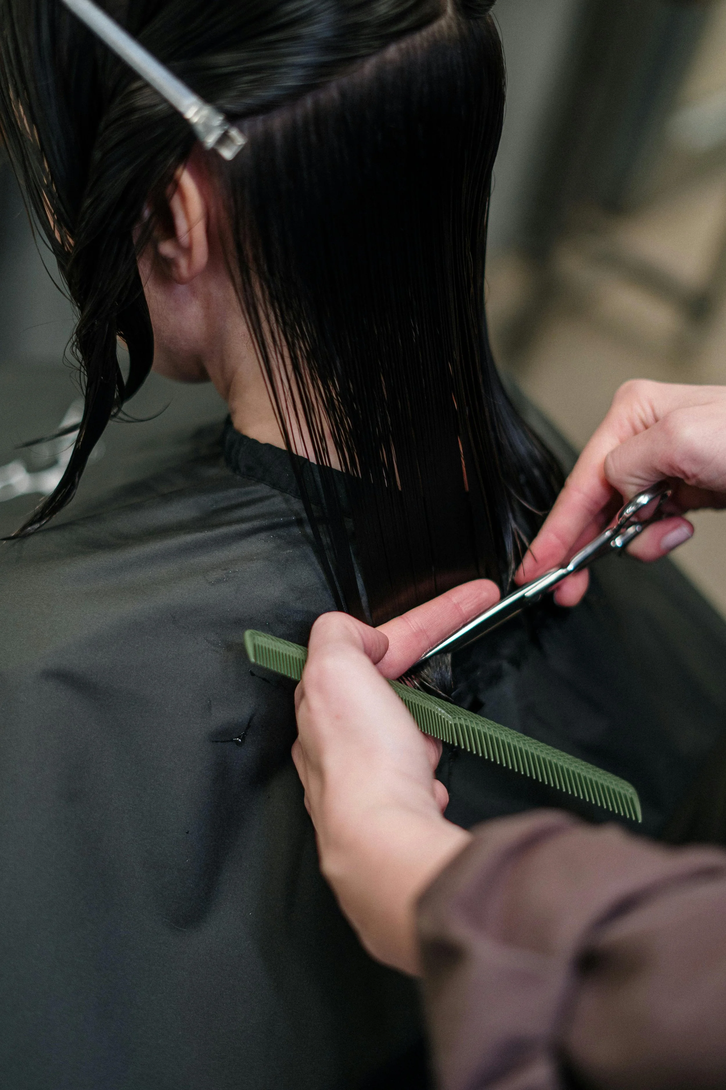 A person getting a haircut, with long dark hair being cut with scissors while sitting in a salon chair, and the stylist using a green comb.