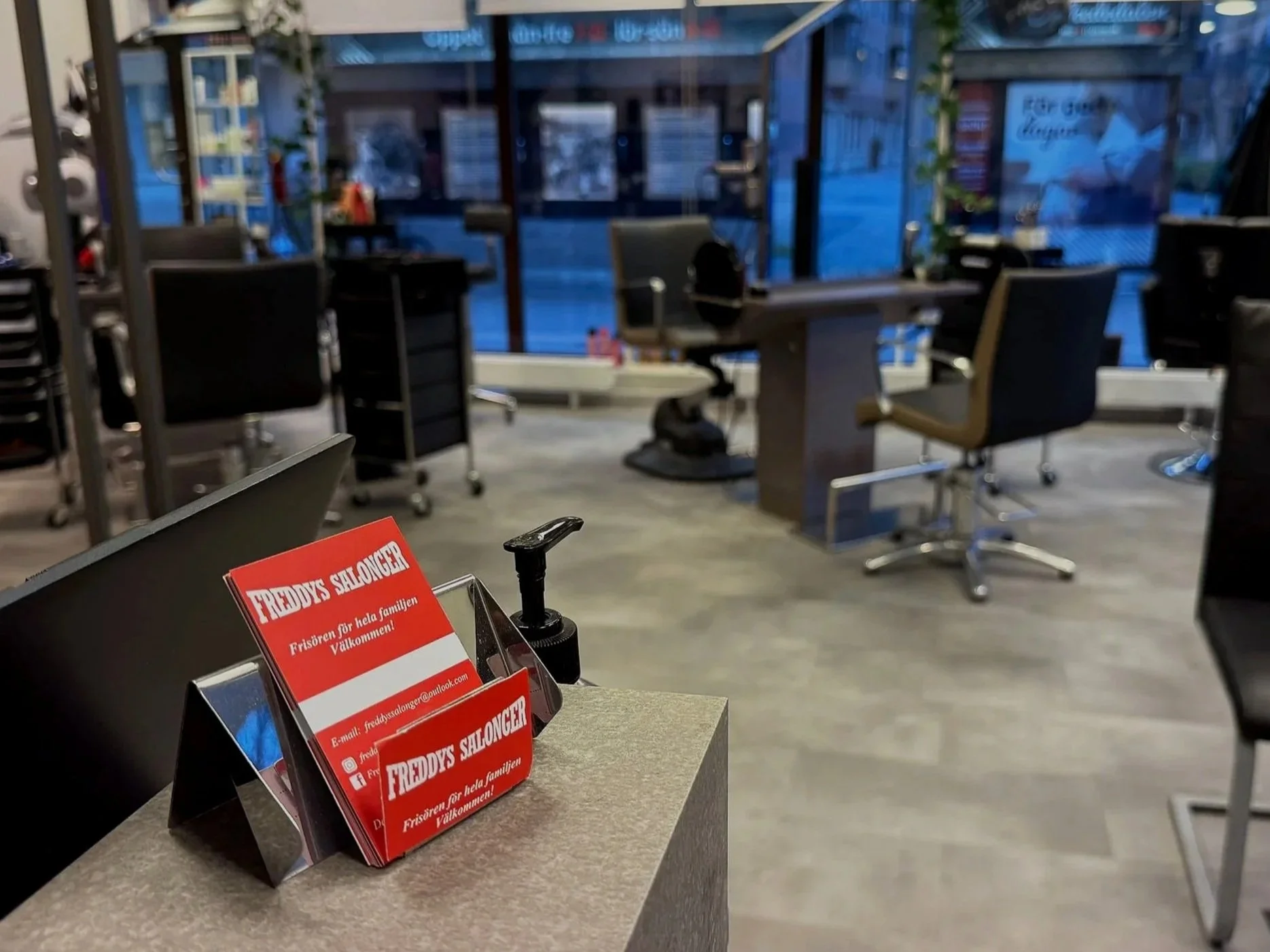 Empty hair salon with chairs, desks, and a red business card holder on a counter in the foreground. Large windows show the street outside at dusk.