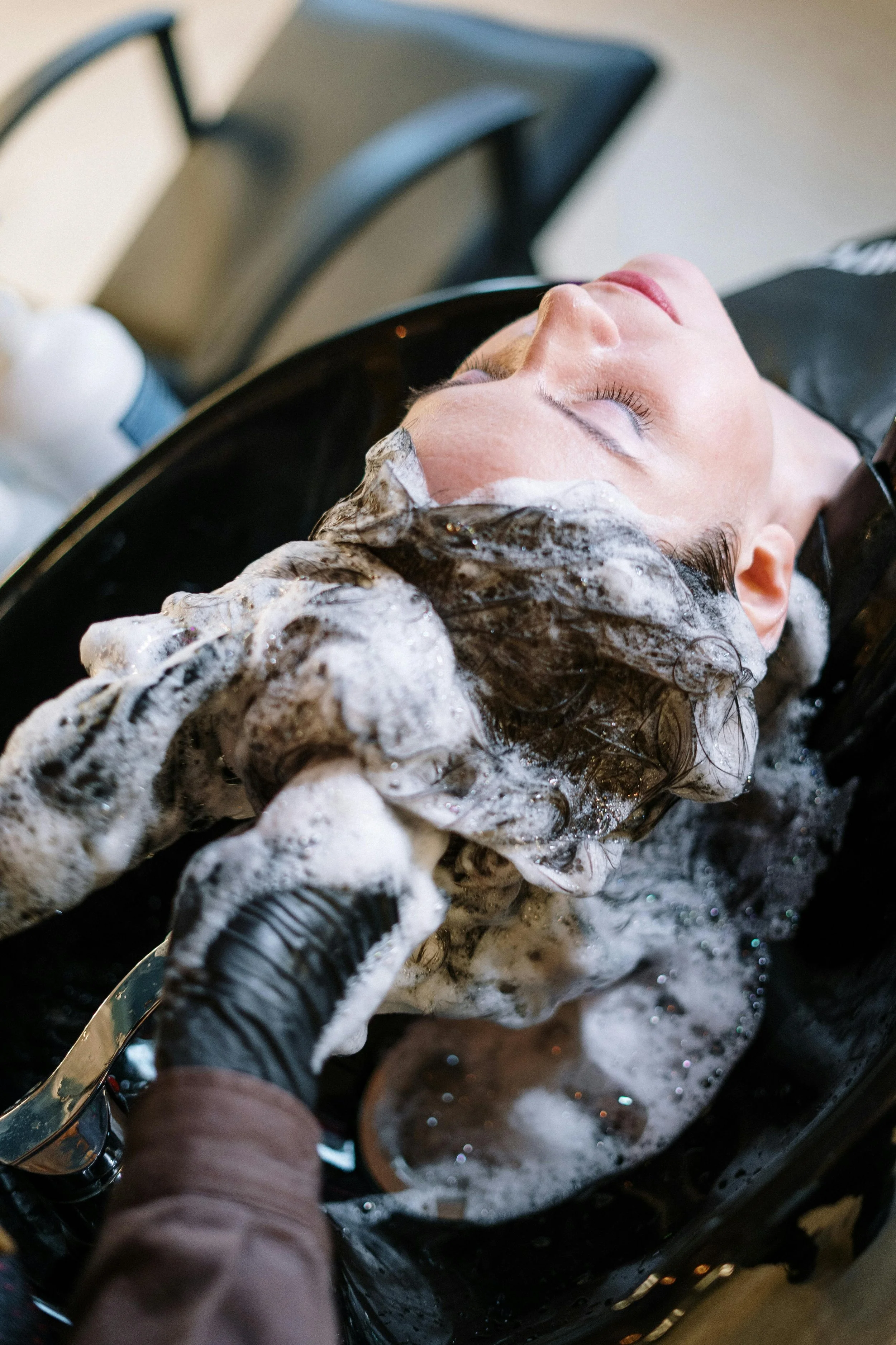 Woman lying with eyes closed at salon sink as stylist washes her hair with soap and water.