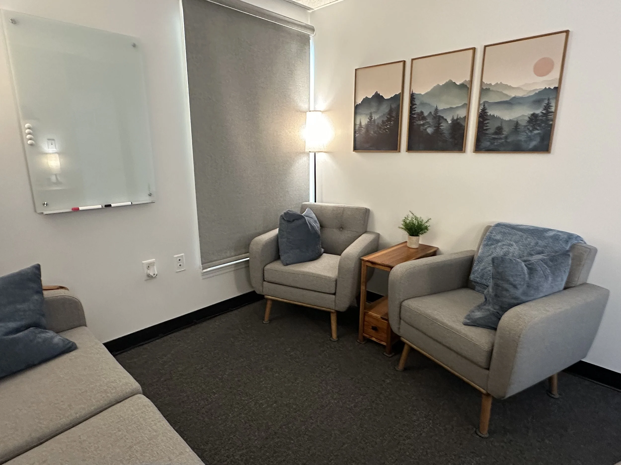 A cozy therapy space for Michael Weiland and Kaleidoscope Counseling with two grey armchairs, a beige sofa, wall art of mountain landscapes, a whiteboard and a window with a grey shade, and a small wooden side table with a potted plant.