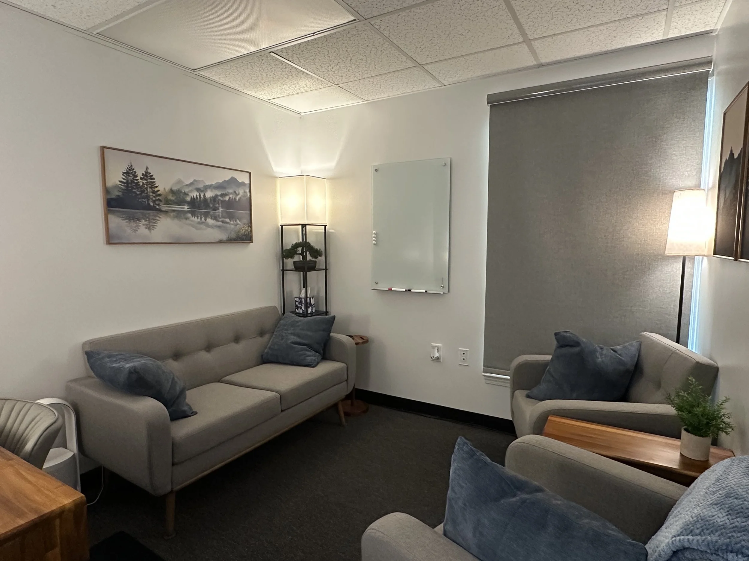 A cozy therapy room for kaleidoscope counseling and Mike Weiland LPC with three gray sofas, blue pillows, a wooden side table, a whiteboard on the wall, and framed landscape paintings, illuminated by a floor lamp and a table lamp.