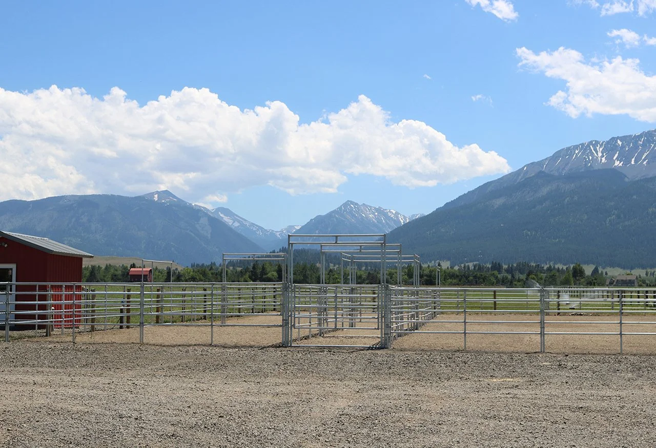 Empty outdoor horse arena with metal fencing and gates, mountains in the background, partly cloudy sky.