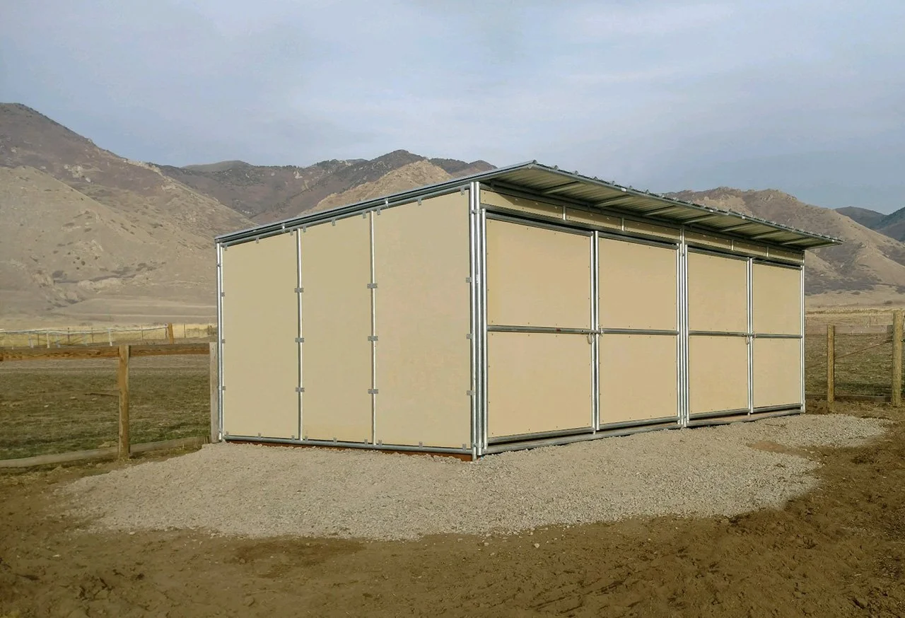 A small beige metal shed with a sloped roof, situated in a rural outdoor area with mountains in the background and a dirt and gravel ground.