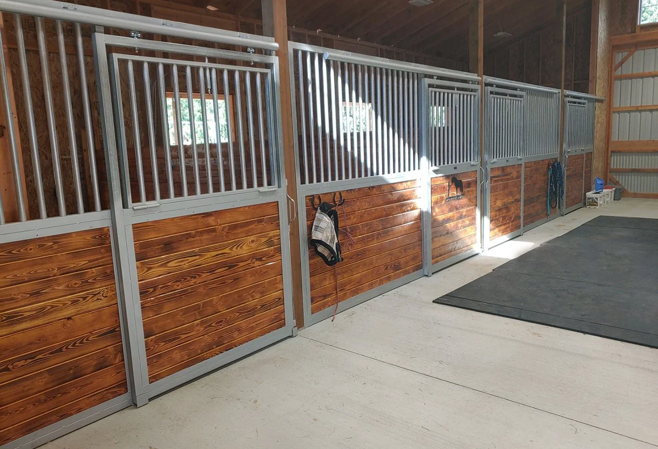 Interior of horse stall barn with multiple stalls, featuring wooden lower panels, metal upper bars, and grooming supplies hanging on the stall doors, with a large black mat on the concrete floor.