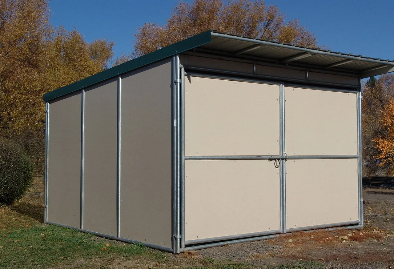 A small, metal shed with a green sloped roof, located outdoors on grass with trees in the background.