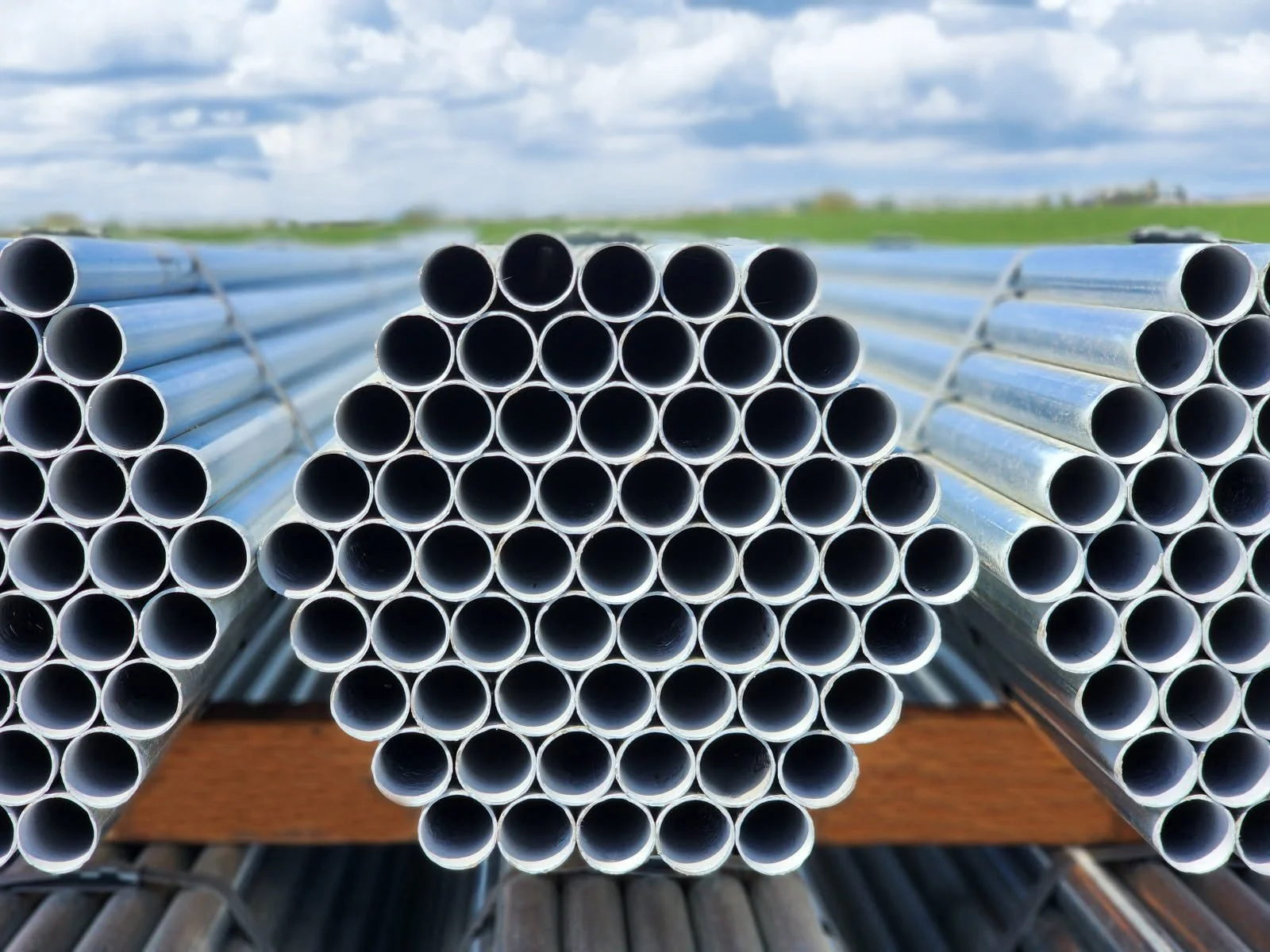 Stack of metal pipes outdoors on wooden beams with a cloudy sky and green grass in the background.