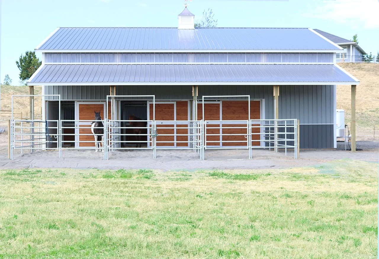 A barn with three horse stalls with two horses inside, surrounded by a grassy field.