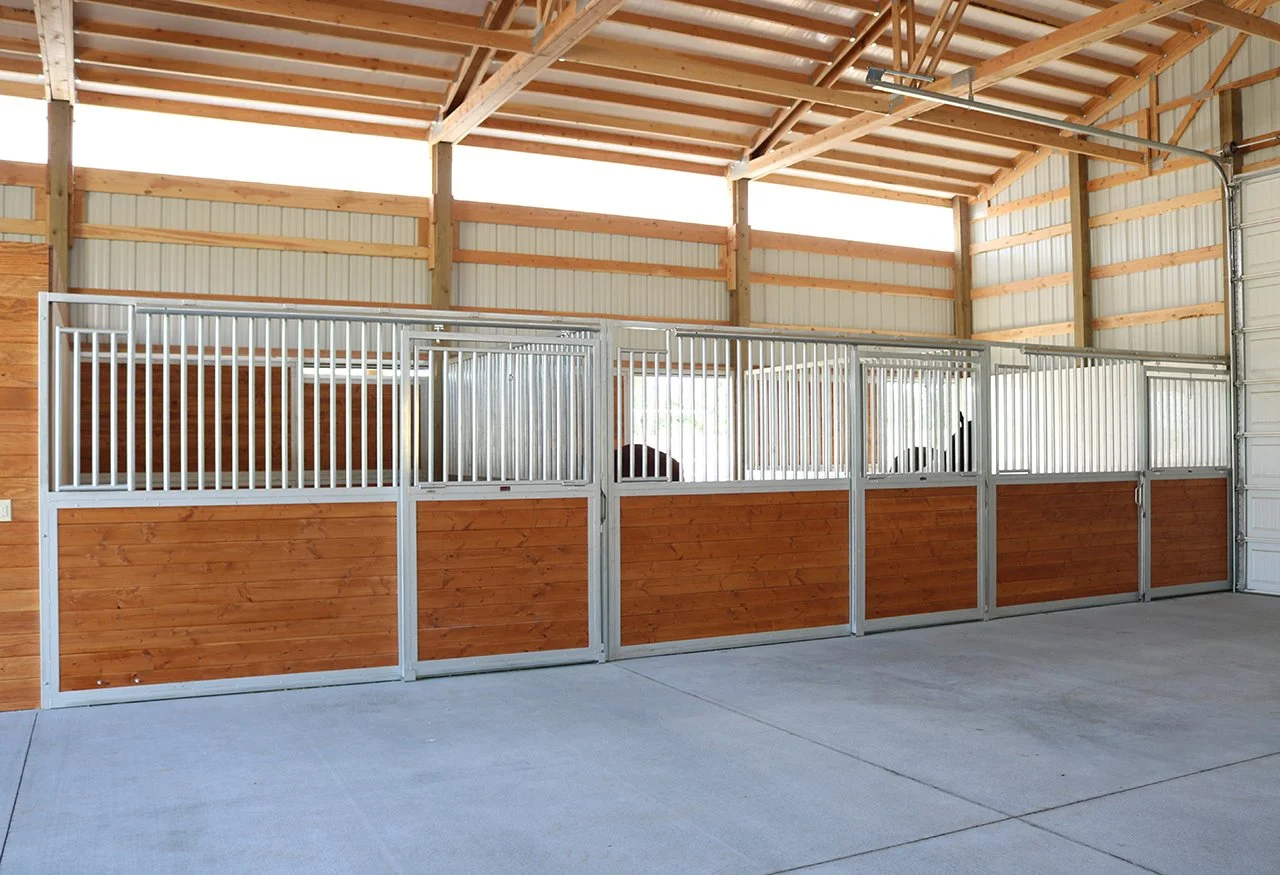 Inside a spacious barn with metal and wood stables, concrete floor, and open ceiling beams.
