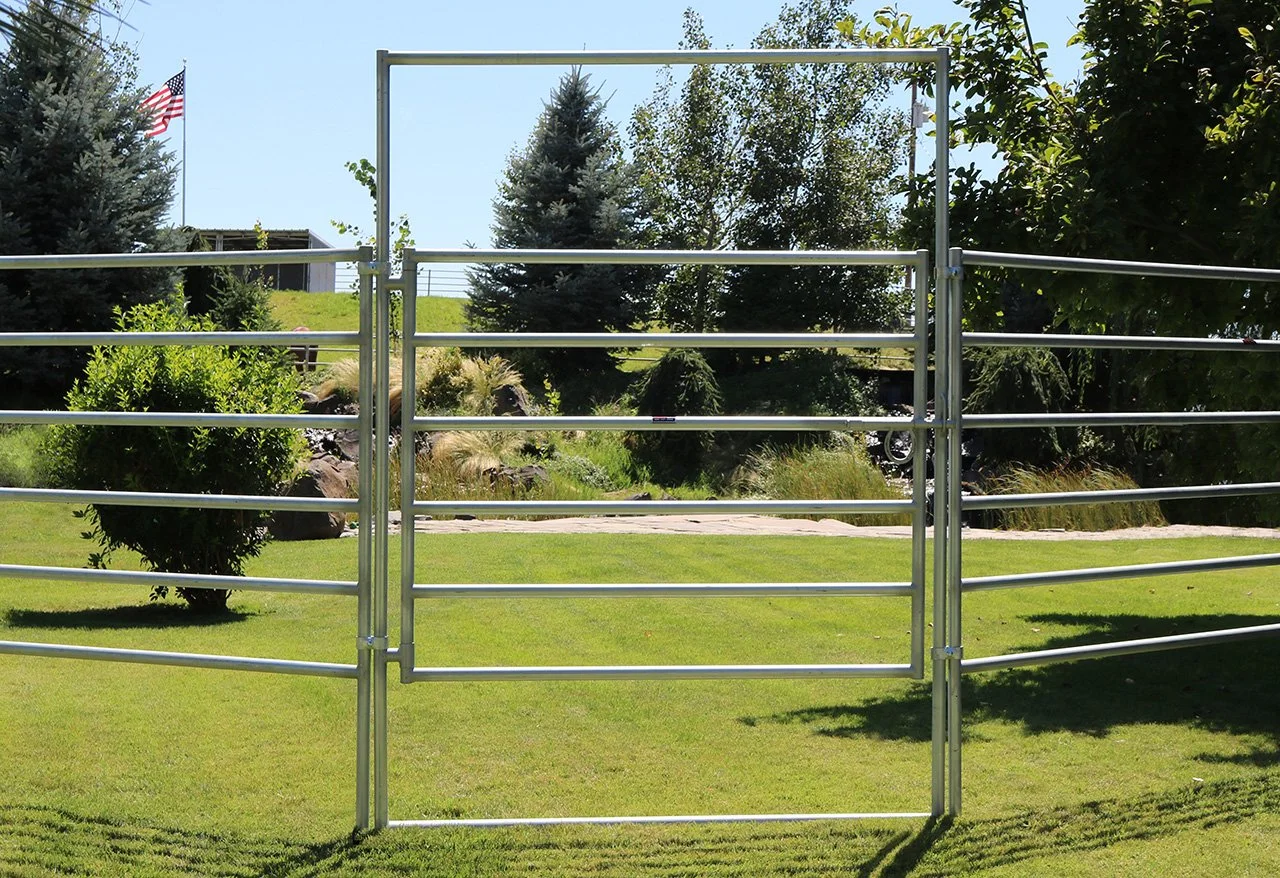 A metal gate in a grassy yard with trees and shrubs in the background, and an American flag flying on a flagpole.