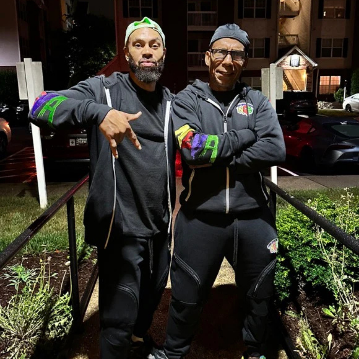 Two men wearing black jackets with colorful arms patches and black pants, standing outside at night, with arms crossed or pointing at each other, in front of a parking lot and apartment complex.