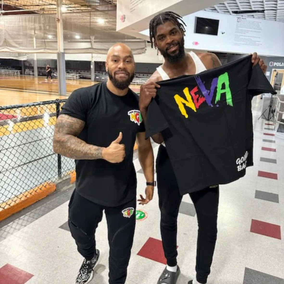 Two men standing in an indoor sports facility, one giving a thumbs up and the other holding a black t-shirt with colorful letters spelling 'N E V A' on it.