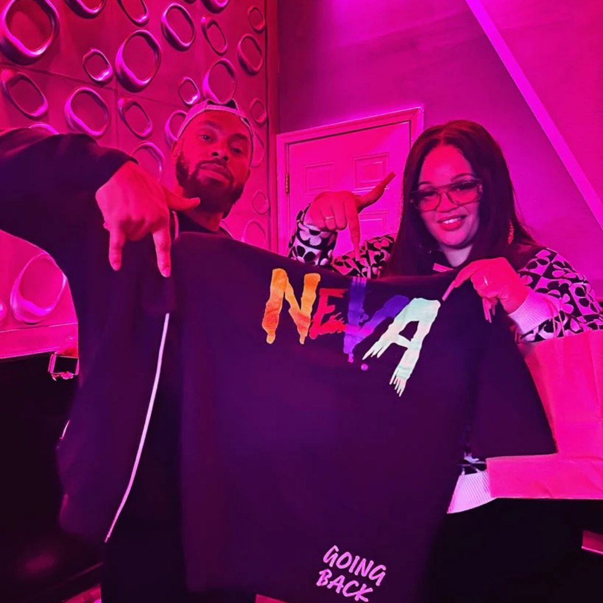 Two people holding a black T-shirt with colorful text, standing in a pink-lit room with patterned wallpaper.