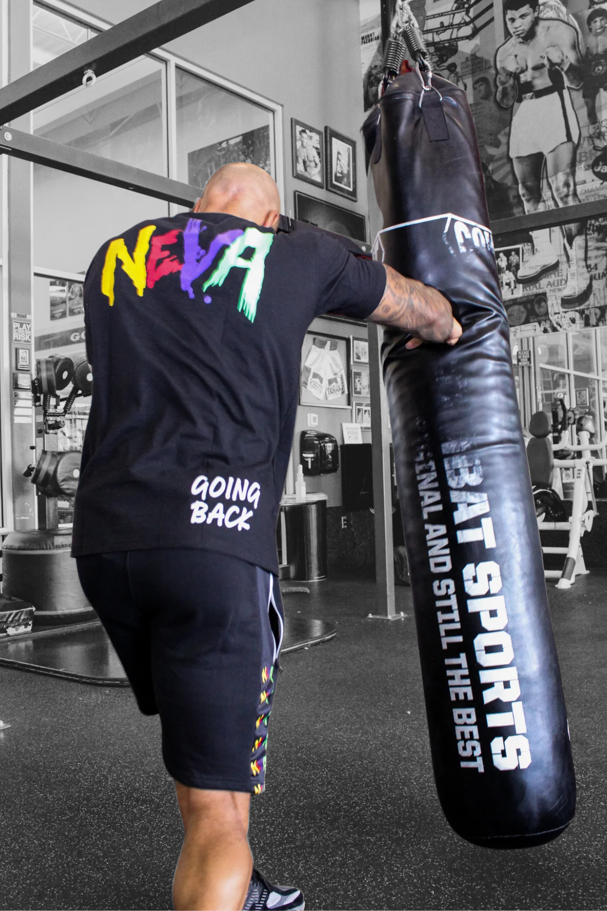 A man with a shaved head and muscular build is practicing boxing with a black punching bag at a gym. He is wearing a black T-shirt with colorful lettering on the back that says 'NEPA' and 'GOING BACK,' and black shorts with colorful designs. The gym has black and white boxing posters and various exercise equipment.