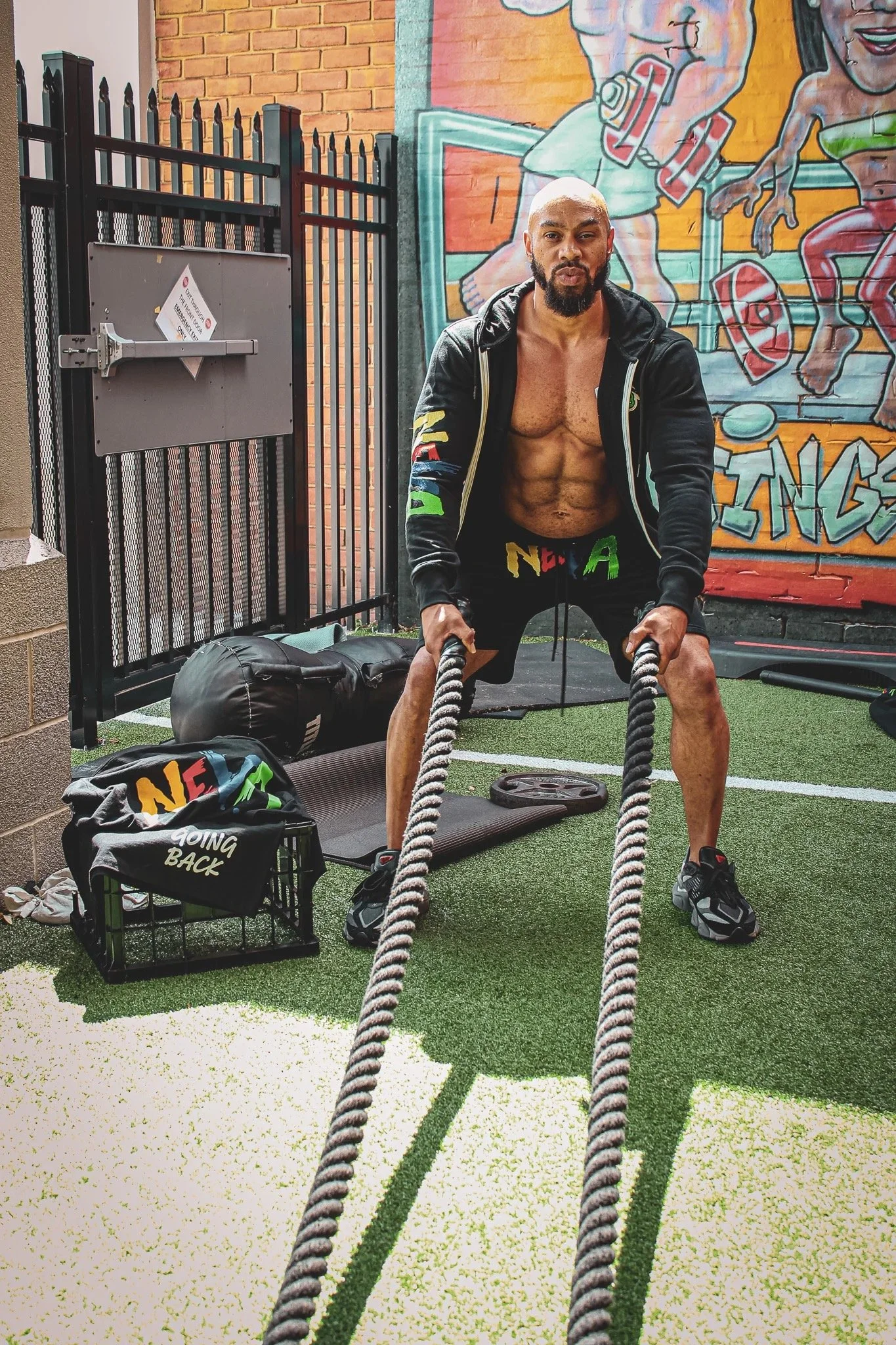 A muscular man with a shaved head and beard, shirtless and wearing black shorts, is exercising with battle ropes in a gym with a colorful mural on the wall.