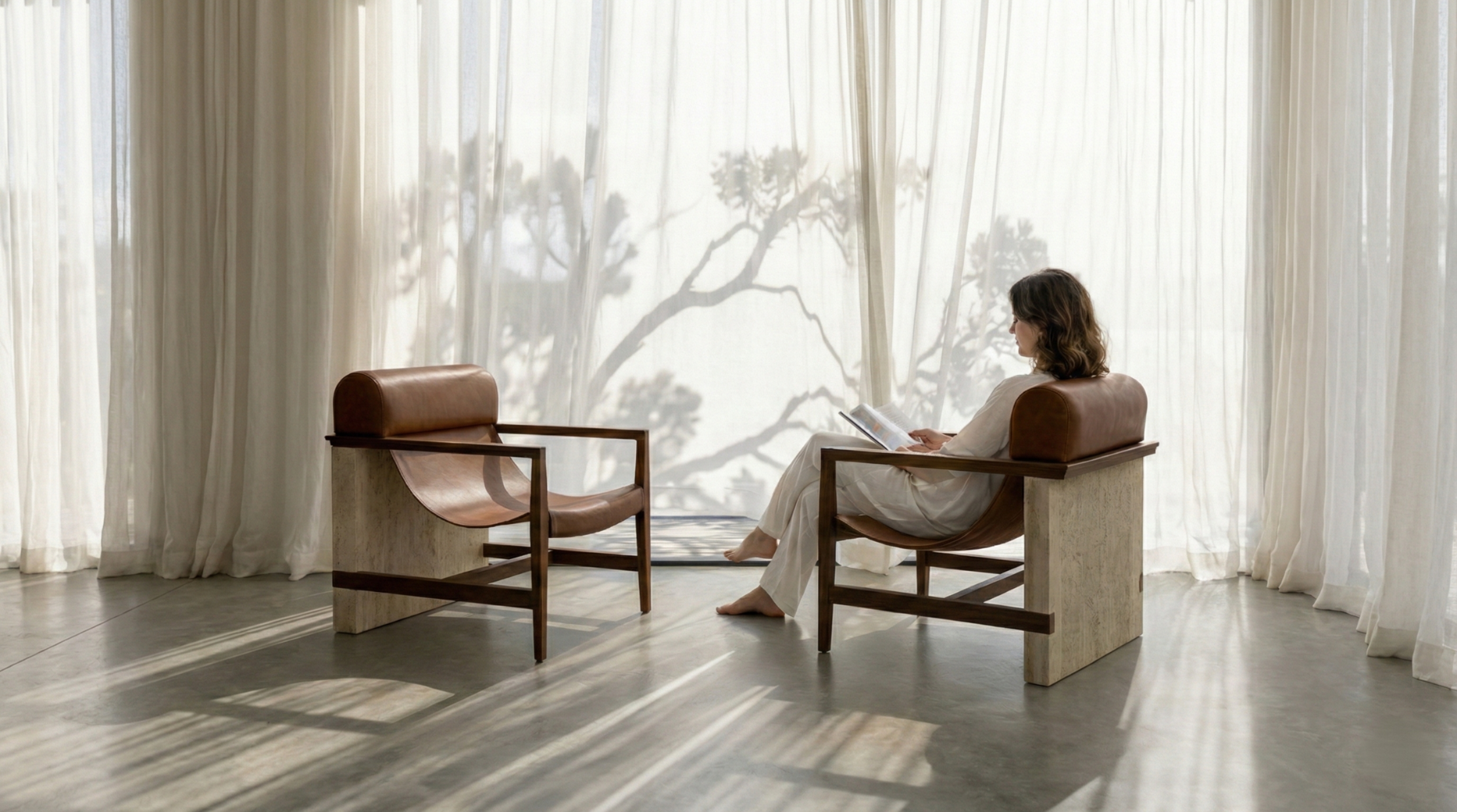 A woman in white clothing sitting on a wooden, stone and leather chair (Punga Chair by Süjä) reading a magazine in a naturally lit room with white curtains and a shadow of a Pohutakawa tree on the curtains and concrete floor.