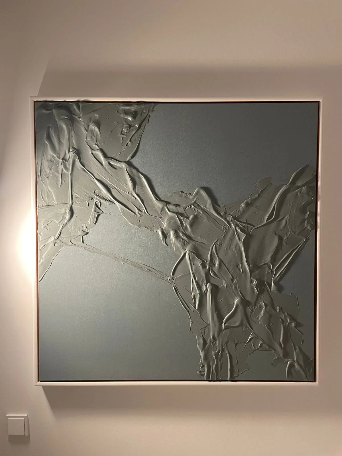 Abstract modern art piece featuring thick, textured silver paint on a square canvas with a neutral background, displayed on a wall with a frame and accent light.