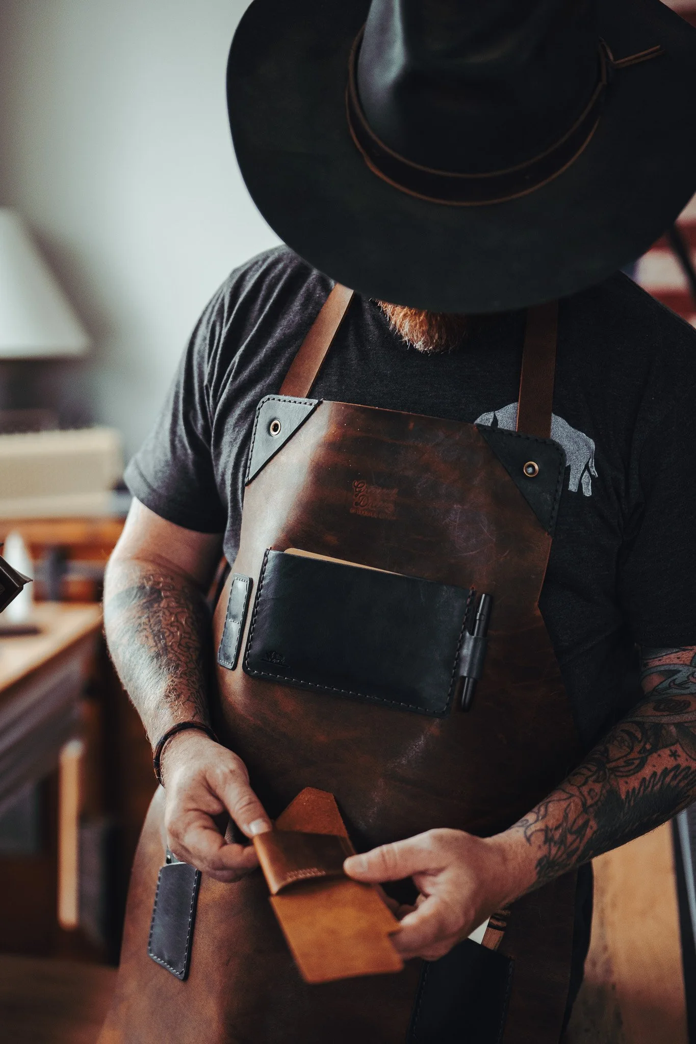Person wearing a black hat, black shirt, and a leather apron, working in a kitchen or barista setting, holding a small cloth or towel.