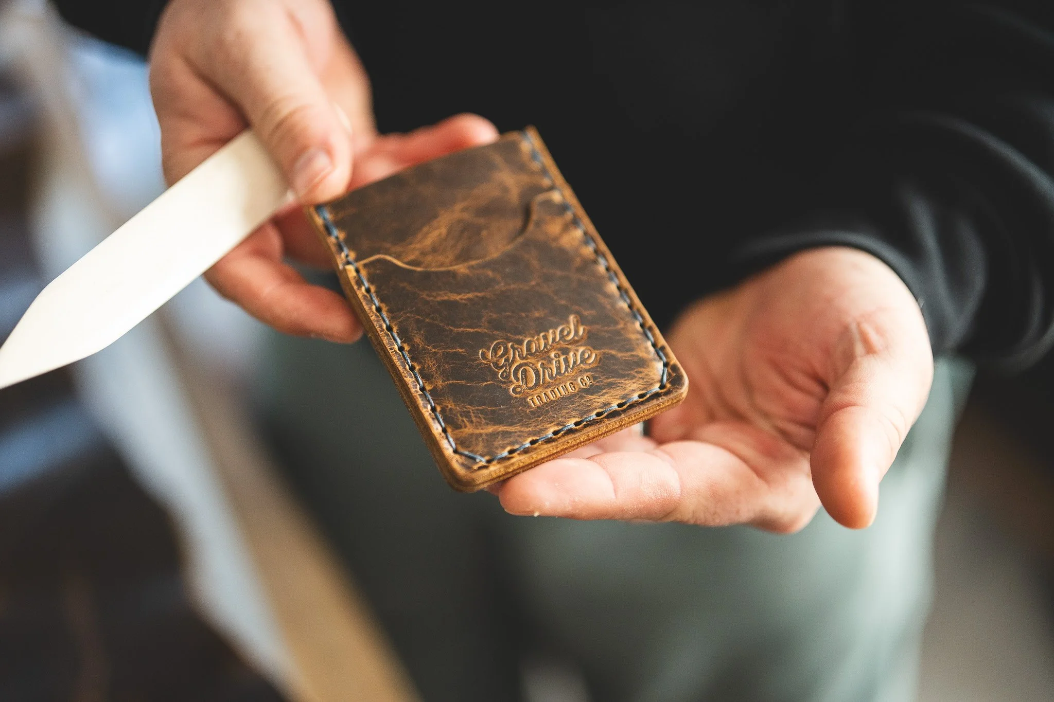 Close-up of a person holding a brown leather wallet with the words 'Gorbel Drive Trading Co.' embossed on it. The person's hand is visible, and they are pulling out or inserting a white card or paper into the wallet.