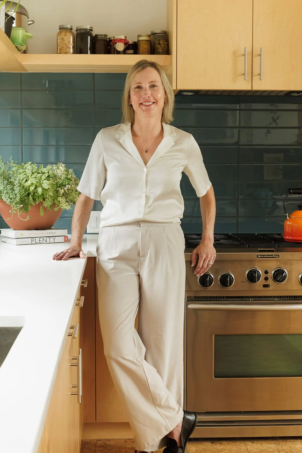 A woman standing in a modern kitchen beside a stove, smiling at the camera. She is wearing a white blouse and beige pants, with a potted plant and a stack of books on the counter nearby.