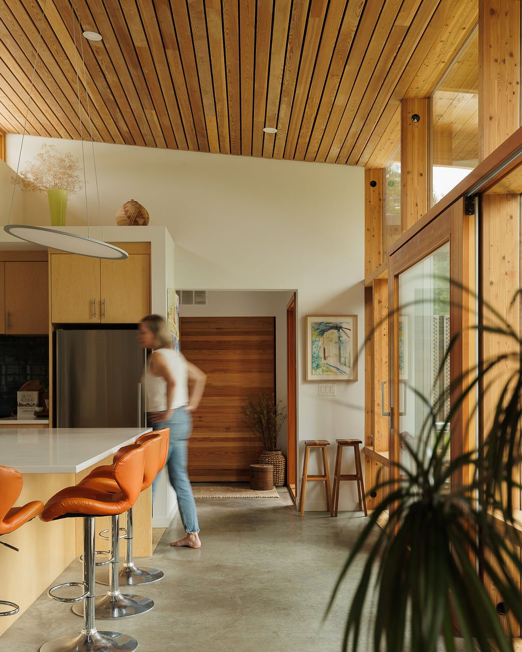 Modern kitchen with wood-paneled ceiling and large windows, featuring orange barstools at a white countertop, a woman in a white tank top and jeans standing near a stainless steel fridge, framed artwork on the wall, and a potted plant in the foreground.